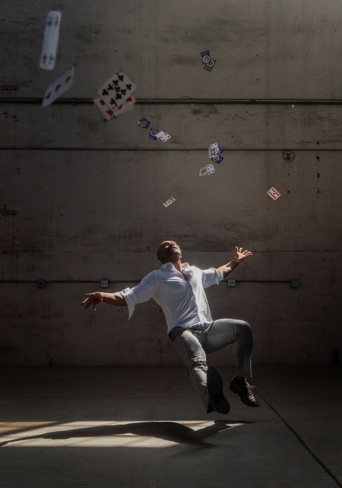 Man in white shirt and grey pants levitating with playing cards floating around him in a dark room.