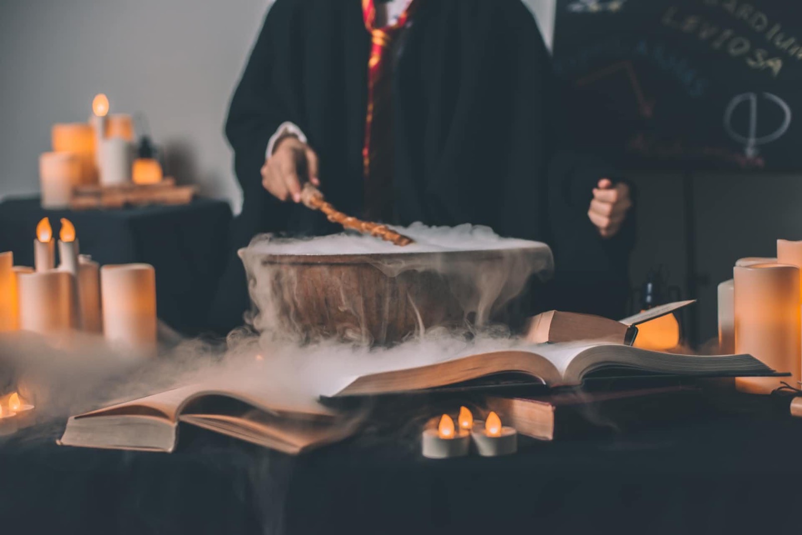 Person in black robe stirring a smoking cauldron with a wand, surrounded by candles and open books.