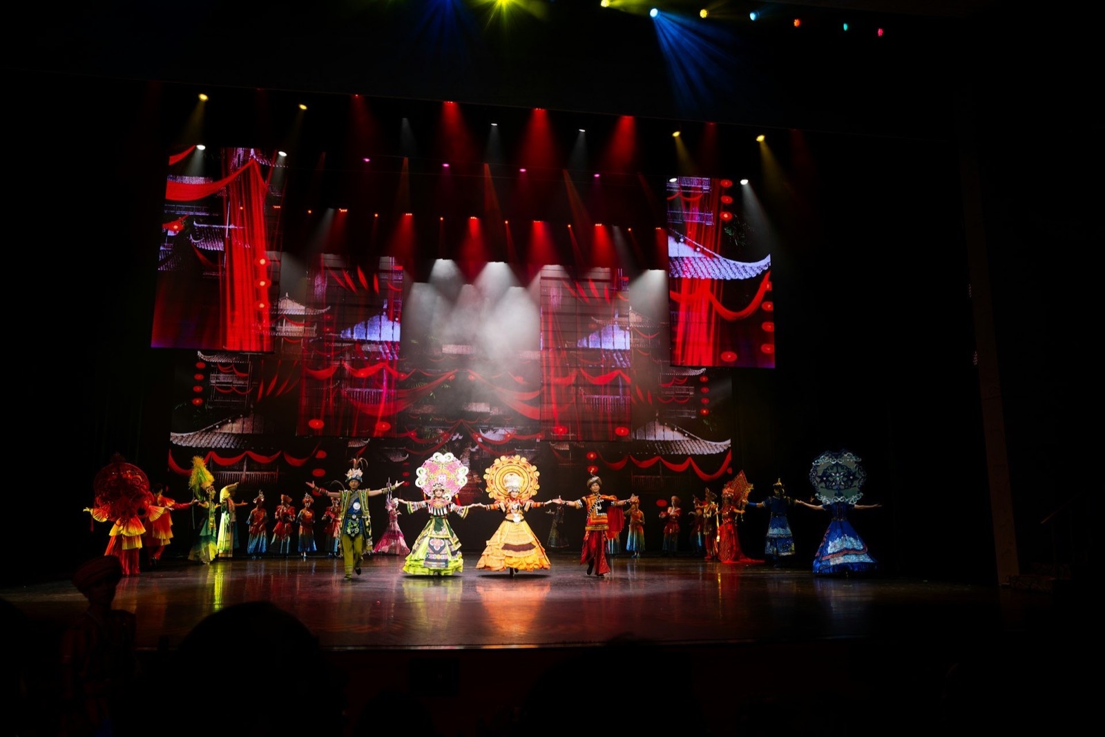 Performers in vibrant, traditional costumes with large, ornate headpieces on a dark stage with red lighting.