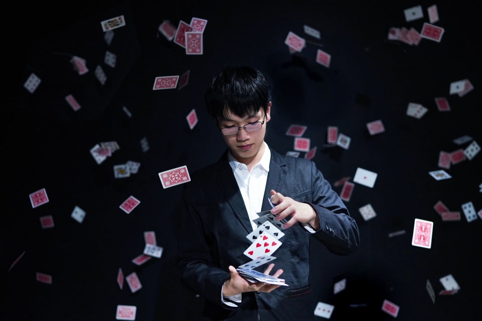 A young man in glasses and a suit performs a card trick with many playing cards floating around him.
